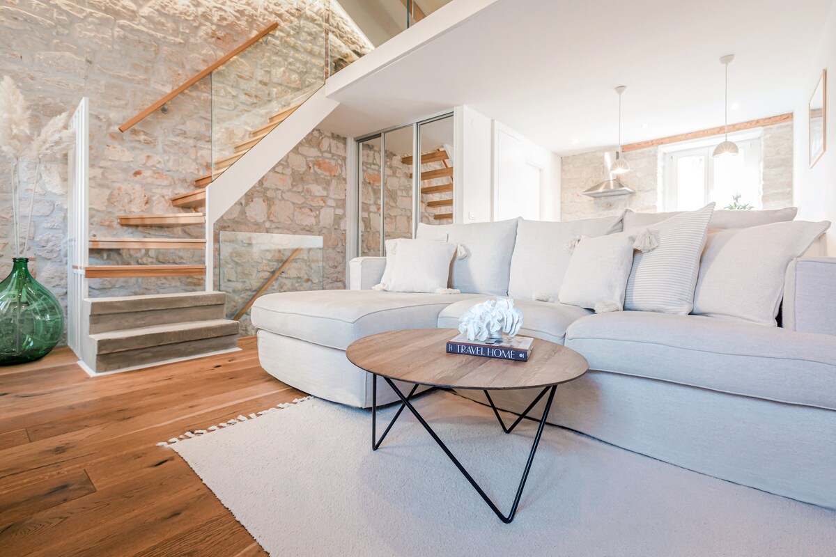 A spacious living area features a light-colored sectional sofa adorned with soft cushions, arranged around a round wooden coffee table. The room's ambiance is enhanced by a textured wall of original stone and a staircase leading to an upper level, illuminated by soft lighting.