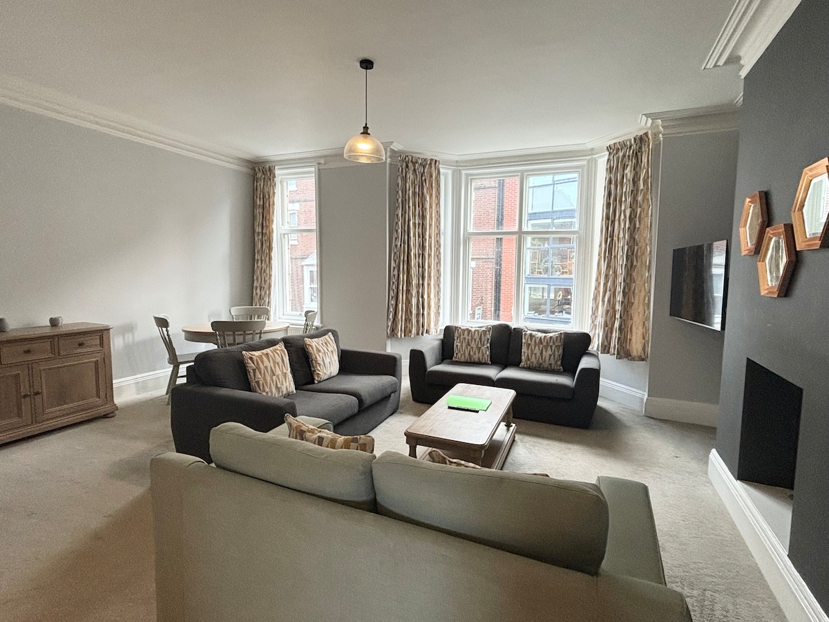 An open plan living and dining area features stylish furnishings, including a comfortable sofa arrangement and a coffee table. Large windows allow ample natural light to fill the space, with soft curtains framing the views. A dining table is positioned nearby, facilitating shared meals.
