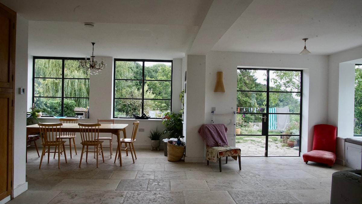 An airy dining area features a large table surrounded by wooden chairs, with large windows offering views of the garden. A cozy red chair is positioned nearby, and natural light illuminates the space. A light fixture hangs from the ceiling, adding subtle charm.