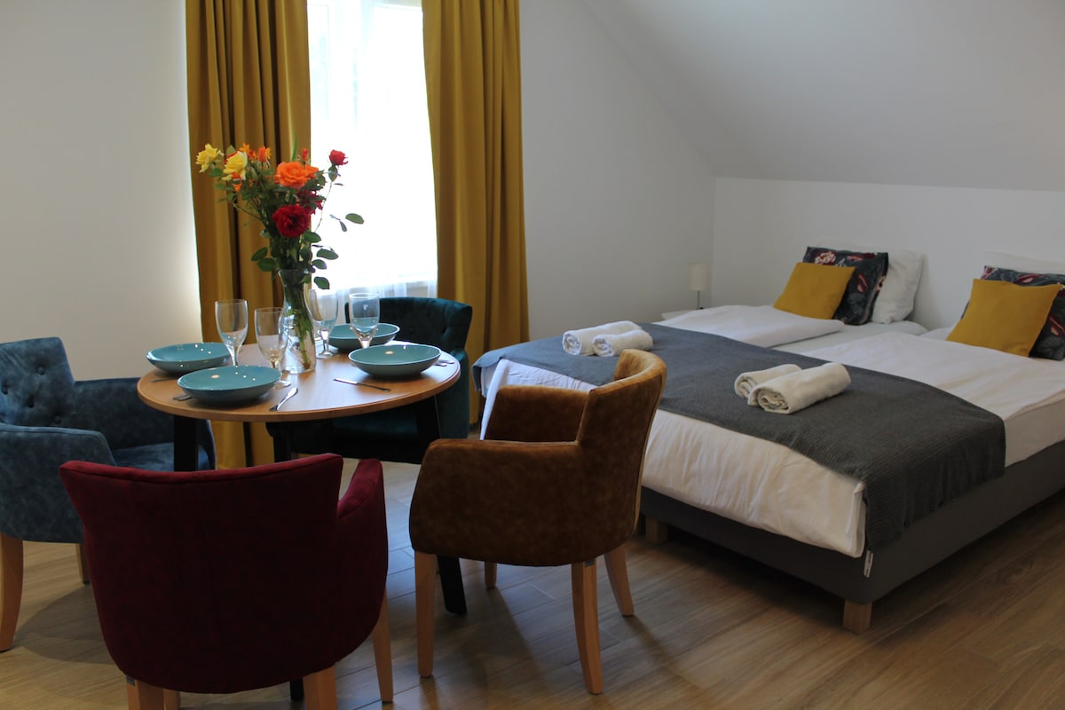 A bright room is presented with a spacious bed dressed in crisp linens, featuring decorative pillows. A small round table, set with dishes and glasses, is surrounded by four chairs in varying colors. Large curtains frame a window allowing natural light to enter.