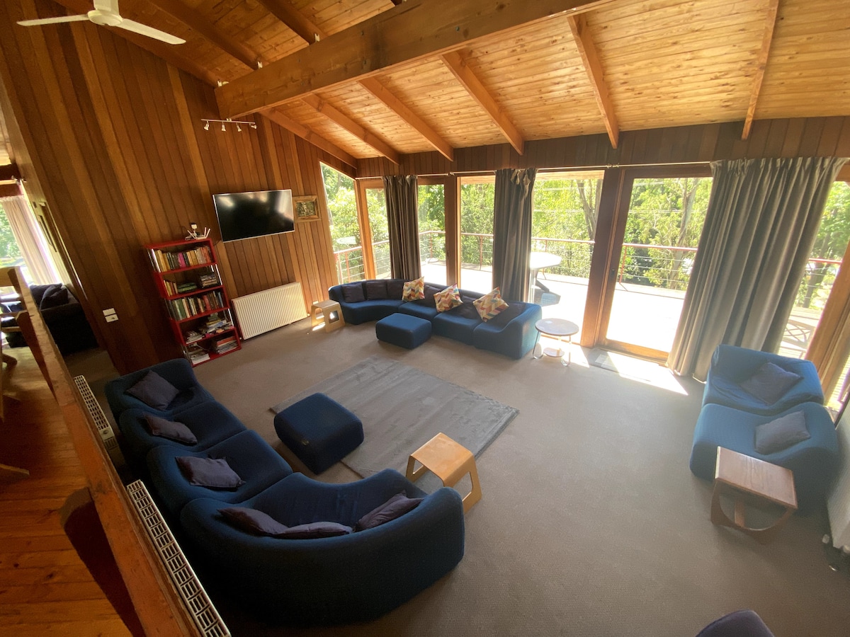 A spacious lounge area features high cathedral ceilings made of timber, offering a cozy atmosphere. Comfortable seating is arranged in a circular layout, complemented by a large rug in the center. Floor-to-ceiling windows provide natural light and views of the outdoors.
