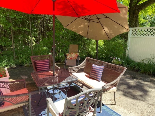 An outdoor seating area is highlighted, featuring two umbrellas providing shade over a set of wicker furniture. The arrangement includes a coffee table, two cushioned chairs, and a sofa, surrounded by greenery and colorful plants along a fence.