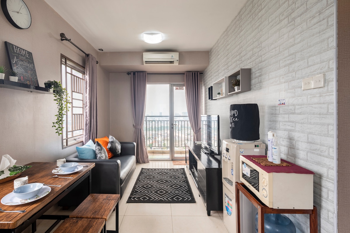 A cozy living area features a modern sofa next to a flat-screen television, with light streaming in through the large window. A dining table is set for four, while decorative elements, such as plants and artwork, add character to the space.
