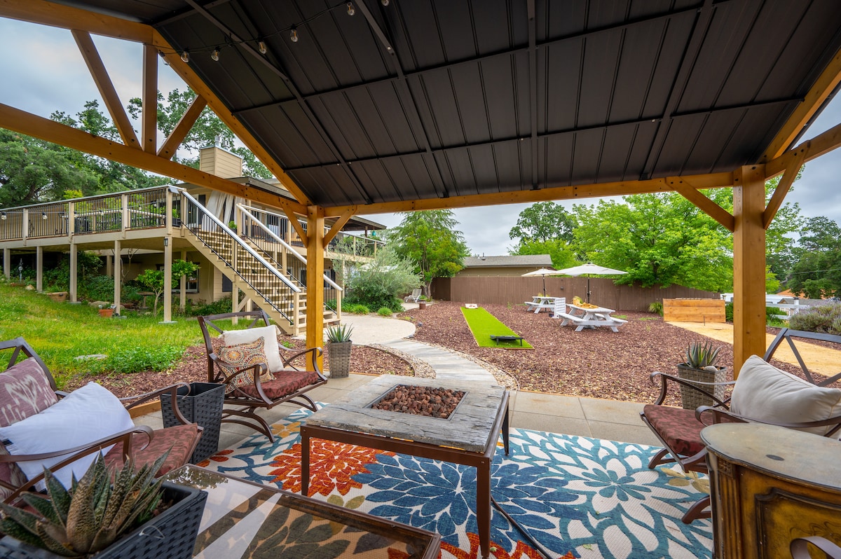 An outdoor seating area features a stone fire pit surrounded by comfortable chairs on a textured rug. A pathway leads to the backyard, where lounge chairs and a green area are visible. Lush trees and landscaping create a calming atmosphere.