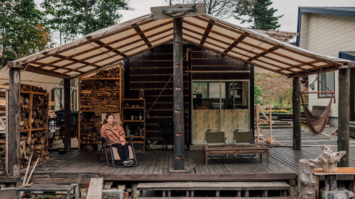 An outdoor deck features a covered area with a wooden structure. Comfortable chairs are arranged around a central space, offering views of the surrounding nature. Several stacks of firewood are stored nearby, and a hammock is visible, providing a relaxed ambiance.