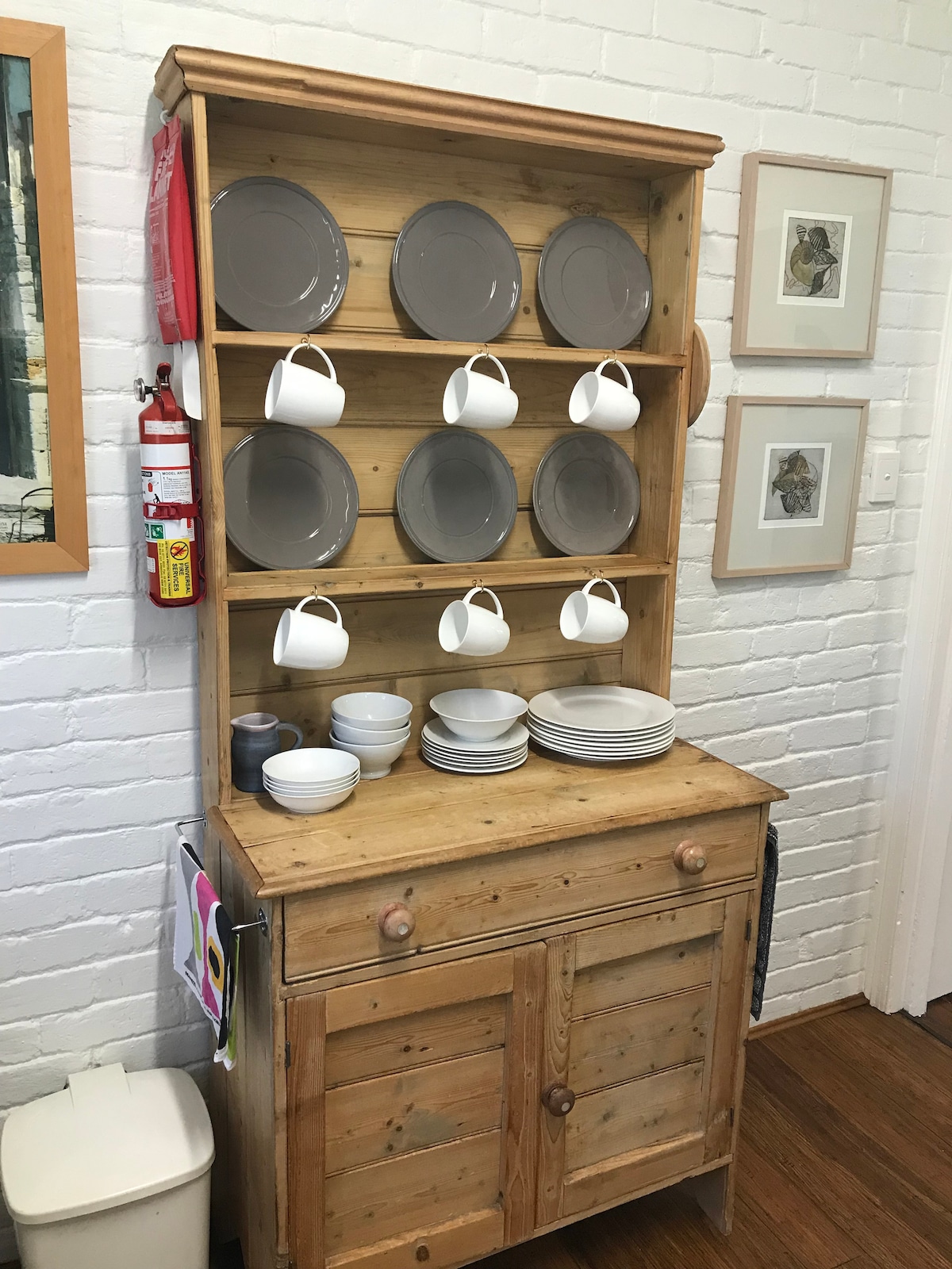 A rustic wooden cabinet is displayed against a white brick wall, featuring several shelves of neatly arranged gray plates and white cups. Stacked bowls and plates sit on the bottom shelf, while a fire extinguisher and framed art pieces provide additional decor.