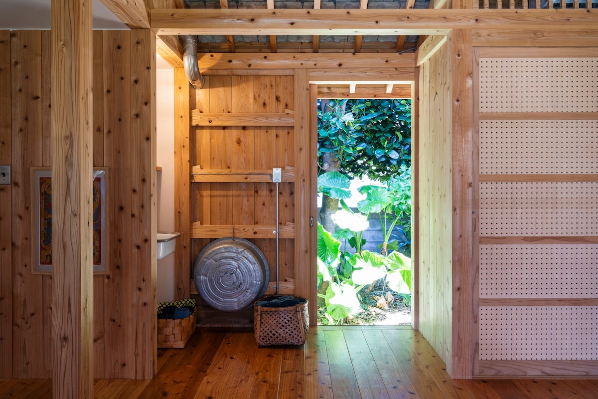 The interior showcases a blend of natural wood textures, highlighted by warm light emanating from an open door. Green foliage is visible just outside, creating a connection with the outdoors. Wooden shelves and baskets add functional decor to the inviting space.