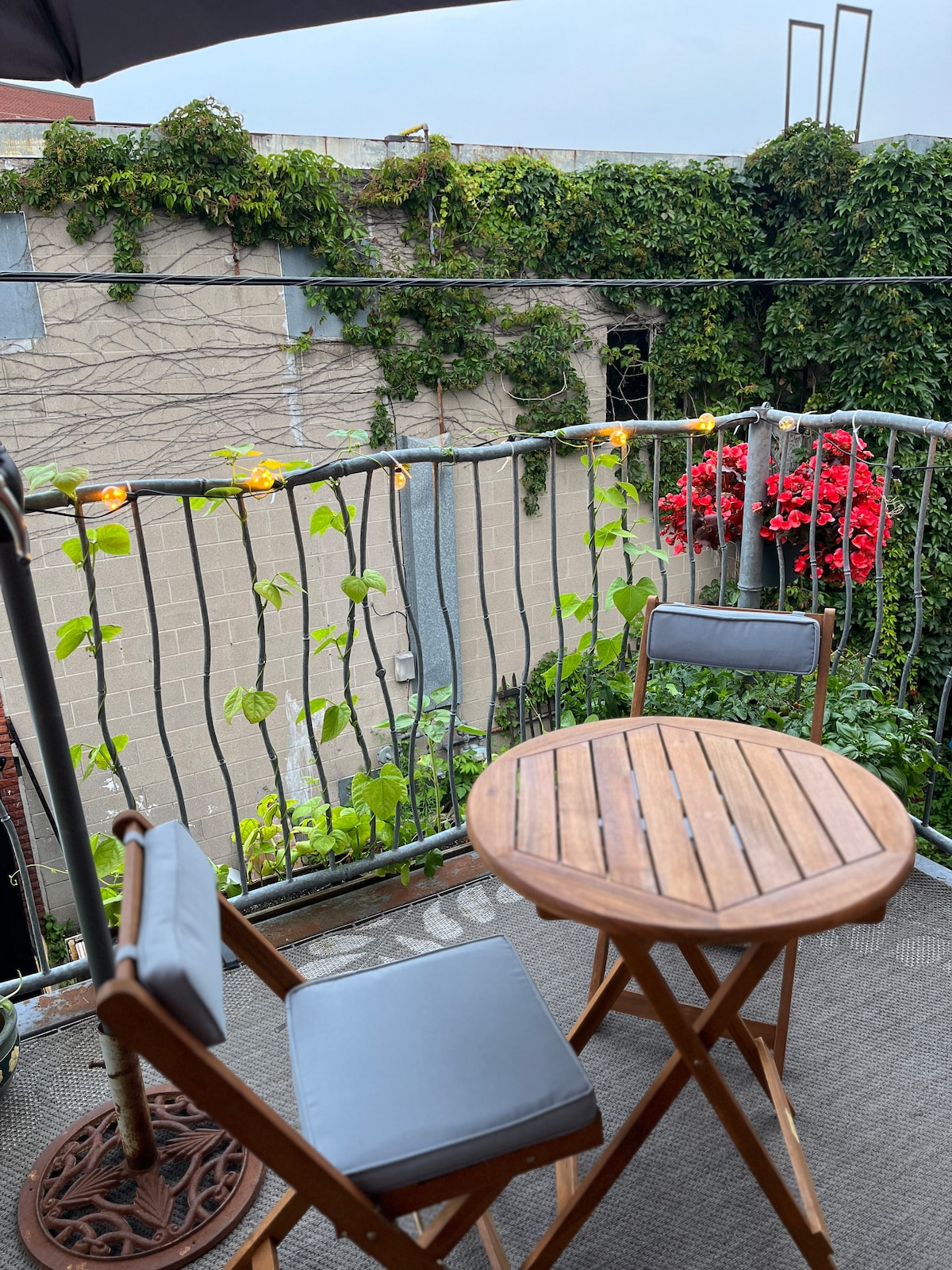 A small balcony features a wooden table and two folding chairs, inviting relaxation. Green vines grow along the railing, with vibrant red flowers adding color. Soft lighting is suspended, creating a calm outdoor space, sheltered by the adjacent buildings.