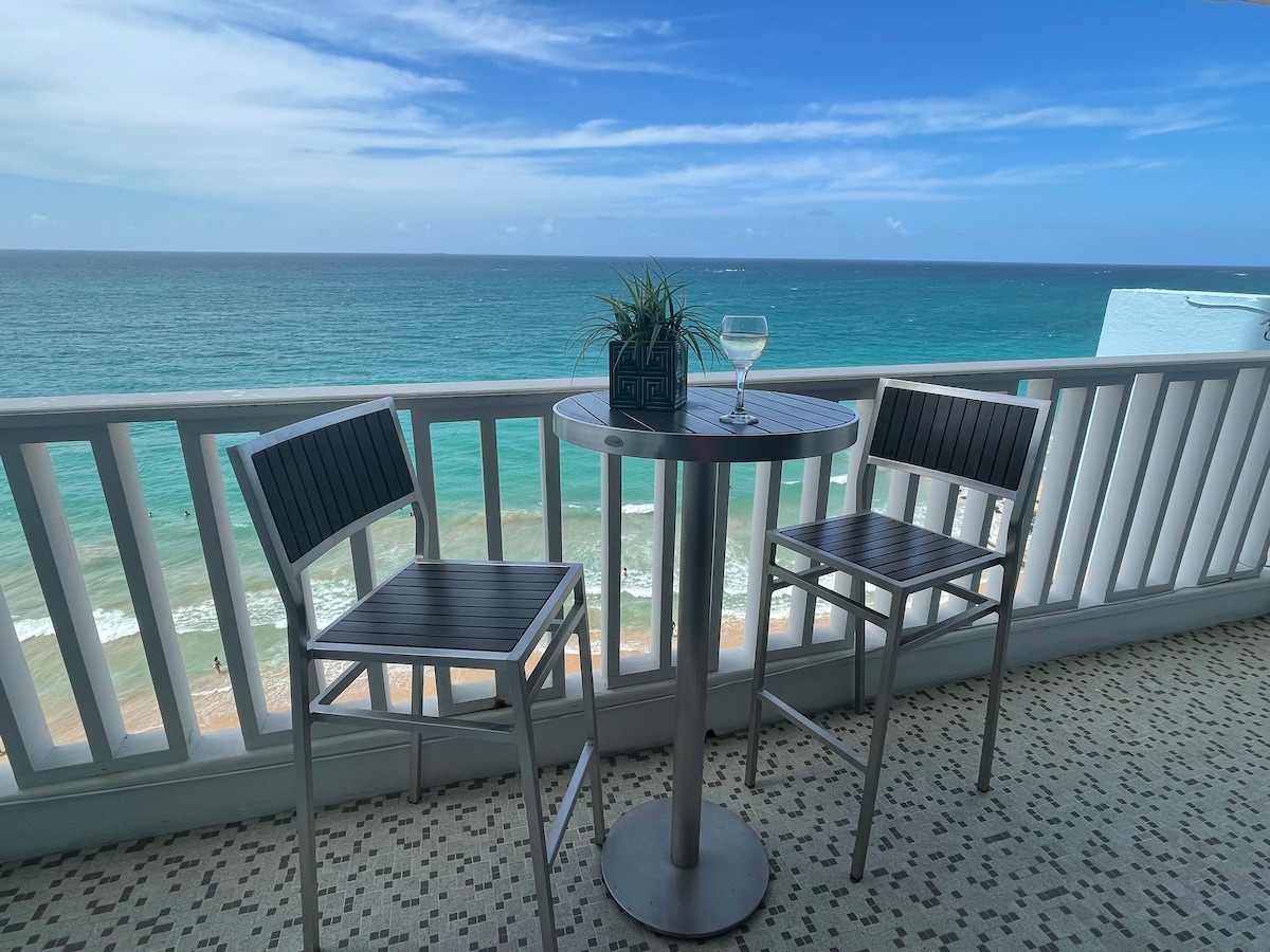 A full-sized balcony features two sleek chairs beside a small round table, offering an unobstructed view of the ocean. A potted plant sits on the table next to a refreshing beverage, with the gentle waves visible in the backdrop under a clear blue sky.