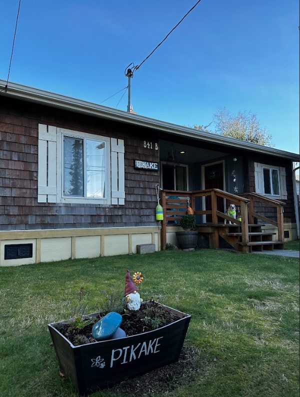 The exterior of the Pikake bungalow features wooden siding and light-colored trim. A welcoming entryway includes steps leading up to the front door. A small garden planter displays colorful decorations, with a gnome and blue object, enhancing the outdoor space. The surrounding lawn is well-maintained.