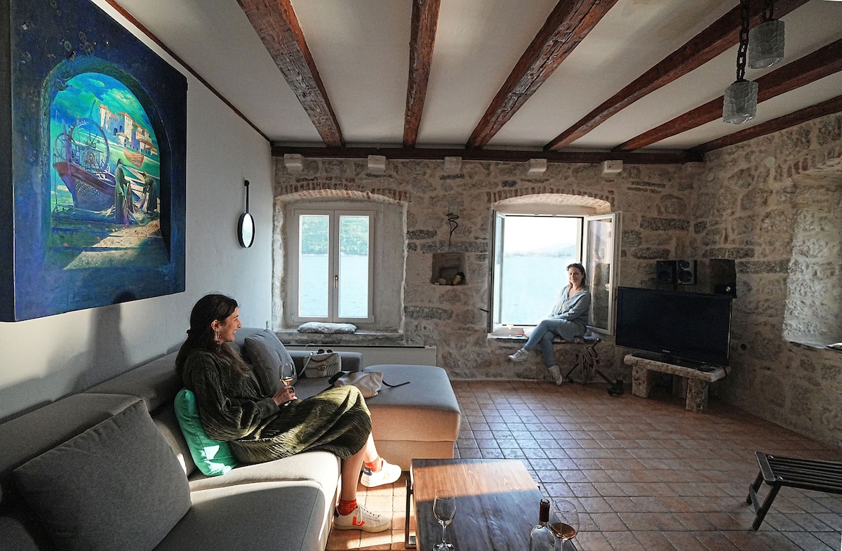 A spacious living area features stone walls and wooden beams, creating a rustic ambiance. A comfortable couch sits nearby, with a view of large windows allowing natural light. A vibrant wall art piece adds color, while a television and a window seat enhance the inviting space.