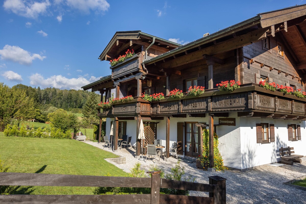 A charming wooden chalet features a two-story design with balconies adorned with flowering plants. A spacious garden area is visible in front, complemented by a gravel pathway and wooden fencing. Lush greenery and trees form a tranquil backdrop under a clear blue sky.