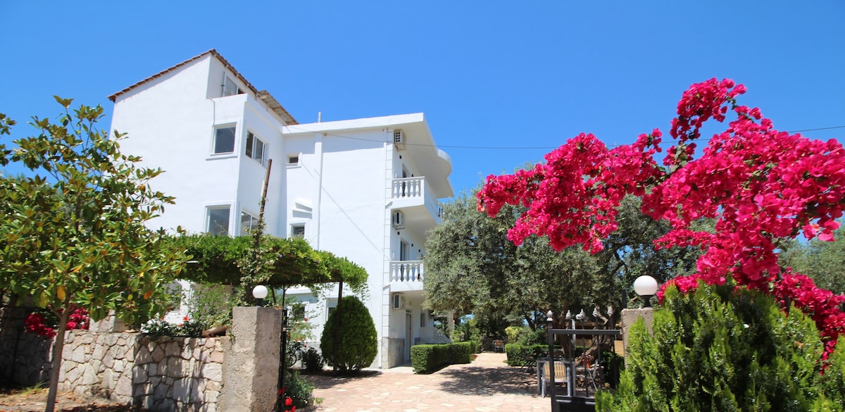 A white three-story villa stands amidst lush greenery, with vibrant pink bougainvillea adorning the entrance. The surrounding garden features well-manicured shrubs and trees, with a stone pathway leading to the building. A clear blue sky provides a bright backdrop to the inviting setting.