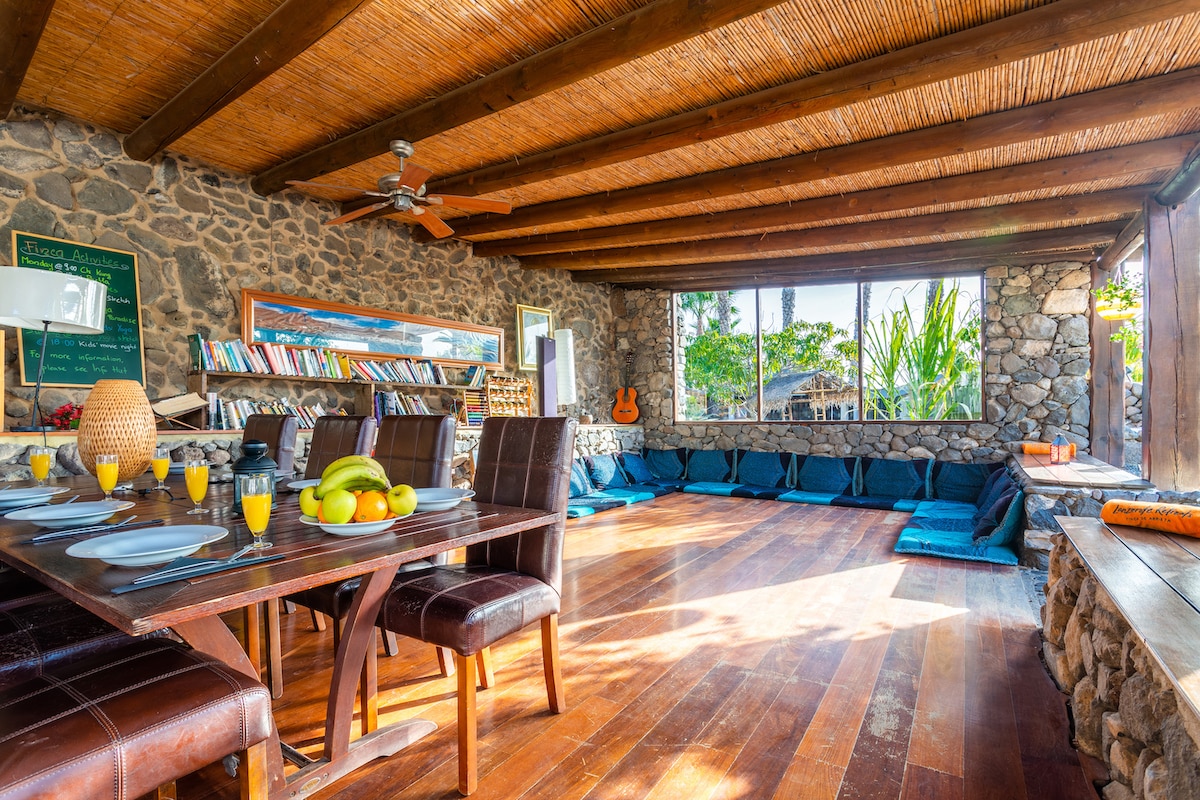 A spacious lounge area features natural stone walls and wooden beams overhead. A dining table is set with dishes and fruit, while a comfortable seating area is arranged with blue cushions. Large windows offer plenty of natural light and views of the surrounding garden.