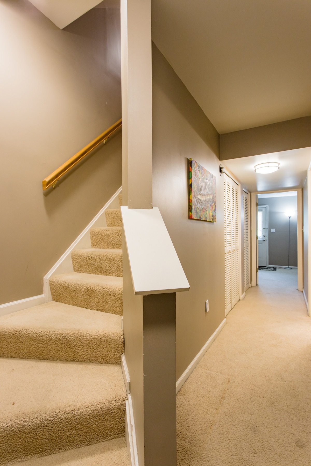 A hallway features soft beige carpeting and neutral wall colors. A staircase ascends to the upper level, flanked by a light fixture overhead. Art hangs on the wall near the stairwell, while two doors are visible at the end of the hall, leading to additional rooms.