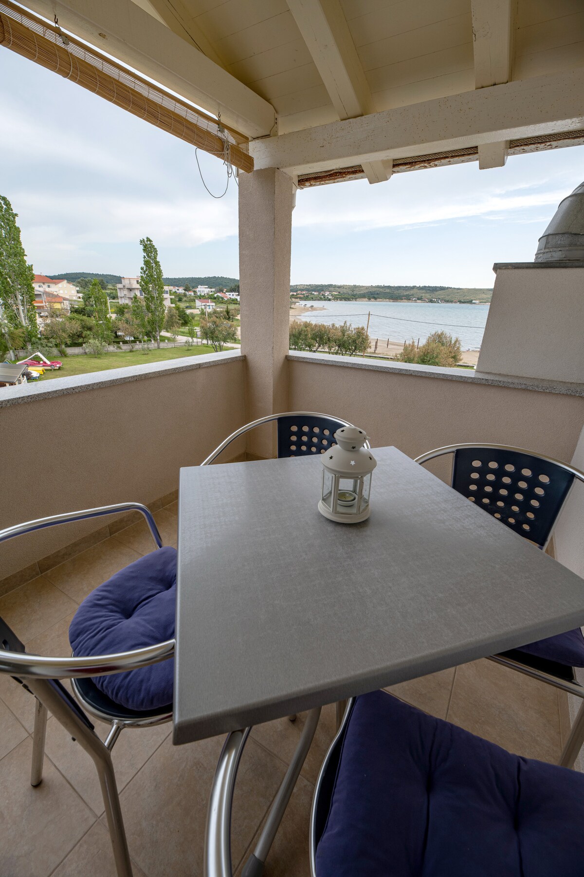 A balcony space features a grey table surrounded by three chairs, with cushions for comfort. A lantern sits on the table, and a view of the sandy beach and serene water is visible in the background, framed by trees and distant hills.