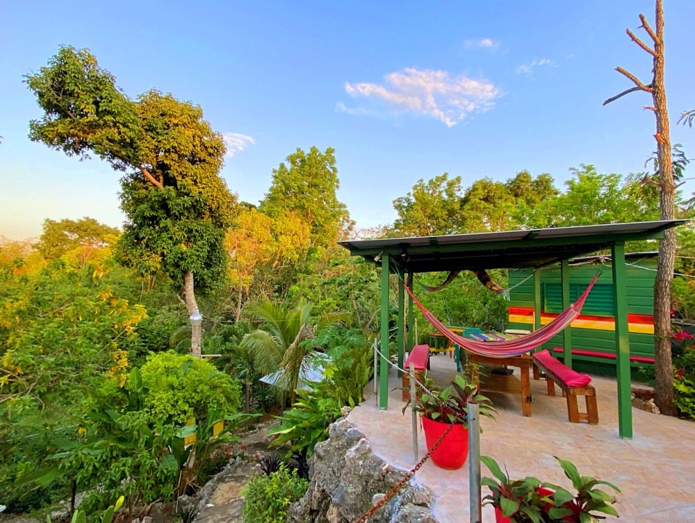 An outdoor lounge area is featured, surrounded by lush tropical greenery. A vibrant hammock is suspended between posts, inviting relaxation. The space includes a wooden table and seating, with colorful walls showcasing local artistry. Sunlight filters through the trees, enhancing the natural ambiance.