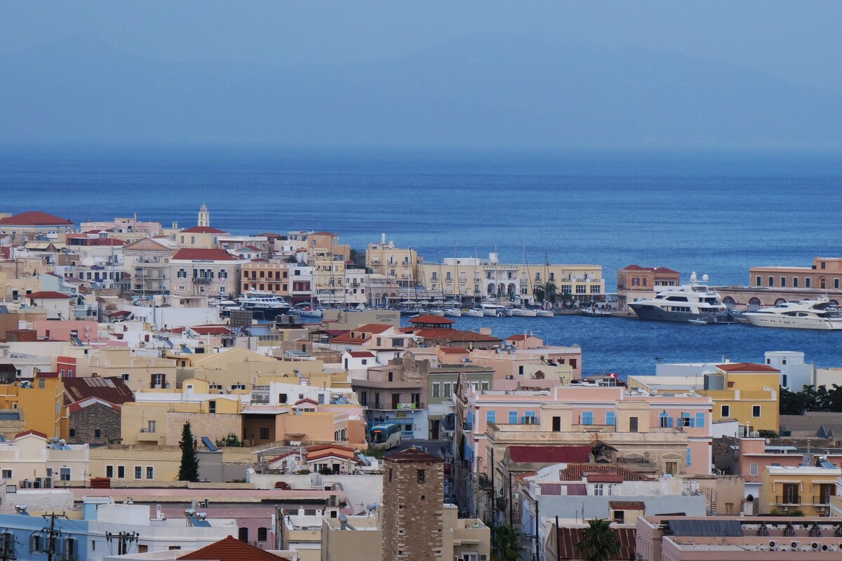 A panoramic view of Ermoupolis is captured, showcasing a coastal landscape with colorful buildings lining the waterfront. The serene blue sea reflects the sky, while distant mountains provide a tranquil backdrop to the bustling town.