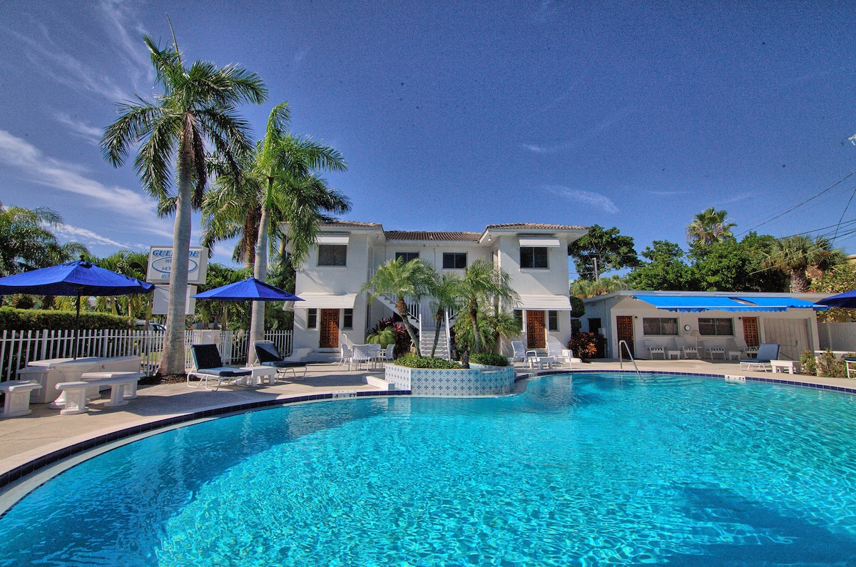 A clear blue swimming pool is surrounded by tropical palm trees and lounging areas with umbrellas. The pool area is bordered by a white fence, with two-story structures visible in the background under a bright sky. Relaxation spaces are offered near the water.
