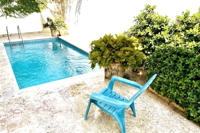 A private pool is surrounded by lush greenery, featuring clear blue water. A single blue chair is positioned beside the pool on a marble-tiled terrace, inviting relaxation. The area is well-lit, with sunlight filtering through the surrounding foliage.
