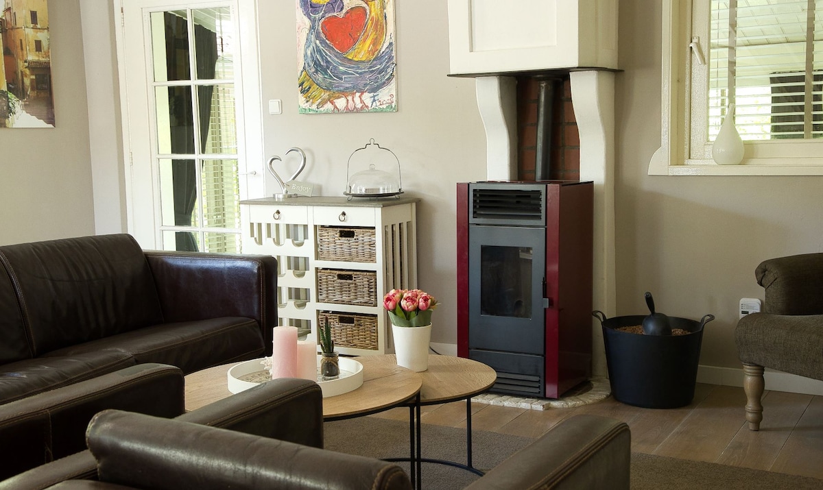 A comfortable seating area features two brown leather sofas and an armchair arranged around a round coffee table. A red pellet stove adds warmth, complemented by a wall with colorful artwork. Light streams through glass doors leading to the outside.
