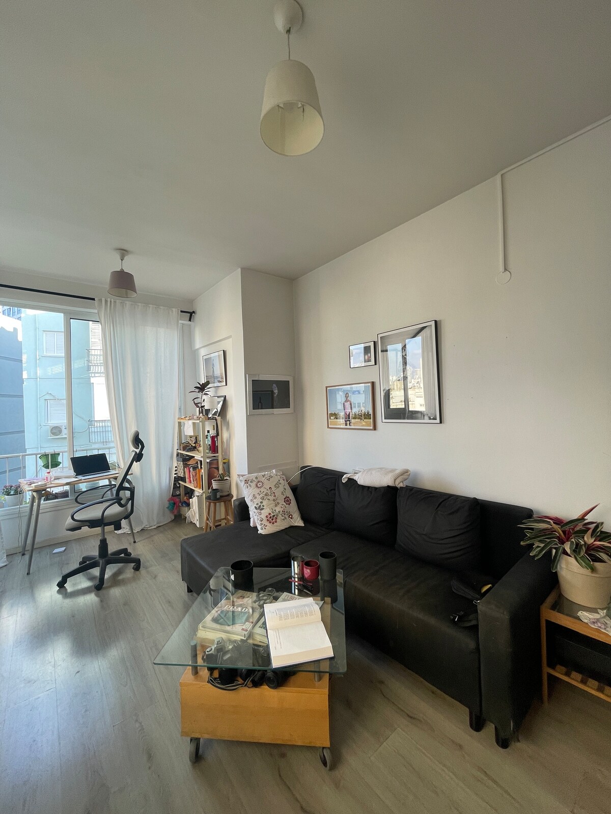 A comfortable living area features a dark sofa adorned with decorative cushions, positioned next to a glass coffee table. A work chair is situated nearby, and wall art adds visual interest. Natural light enters through a large window, complemented by sheer white curtains.