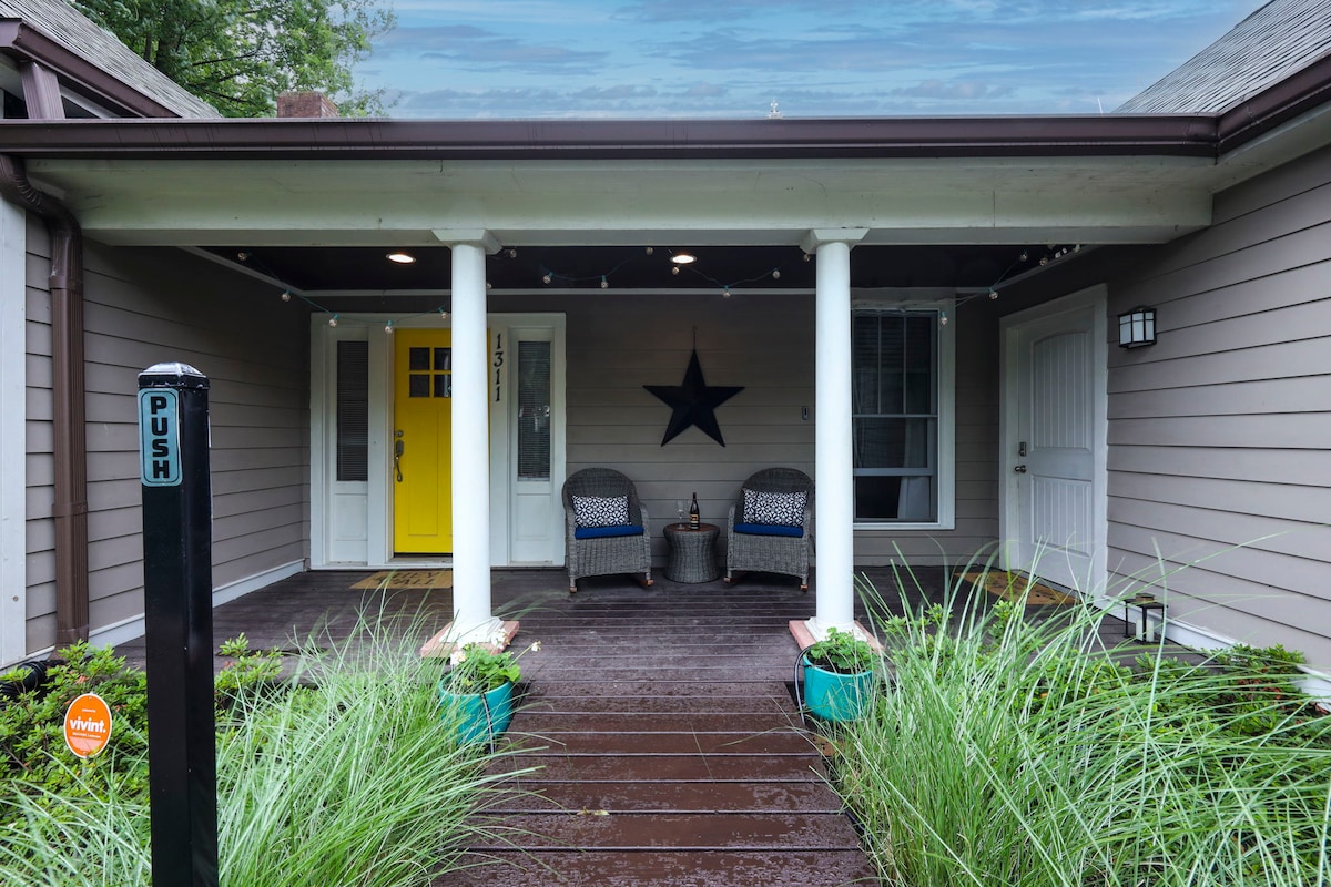The front porch features two comfortable wicker chairs placed beside a small table, accompanied by decorative plants in blue pots. A bright yellow door serves as an inviting focal point, while two white columns support the roof, enhancing the cozy entrance.