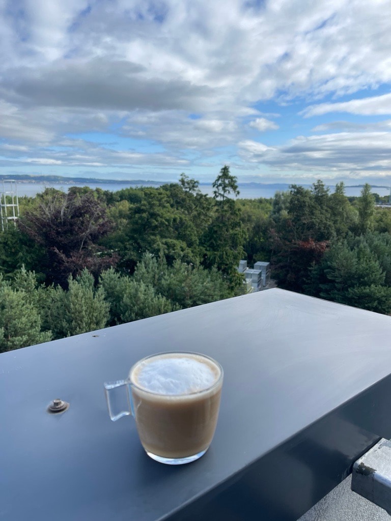 A clear view is captured from the balcony, showcasing a lush landscape of trees under a partly cloudy sky. In the foreground, a glass of coffee with frothy milk sits on a smooth, dark tabletop, enhancing the inviting atmosphere.