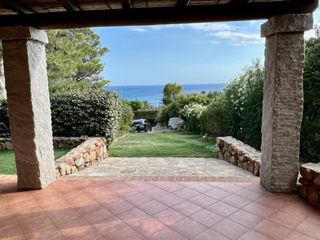 A covered terrace is shown, featuring textured stone columns and a tiled floor. Beyond the terrace, lush greenery leads to a view of the sea, with the tranquil blue water visible in the distance under a clear sky.