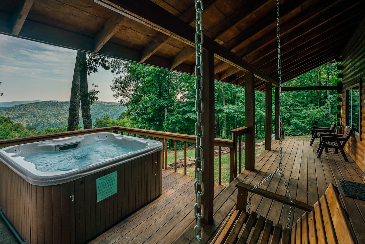 An outdoor deck features a hot tub, accompanied by wooden swings. The expansive view of the surrounding trees and mountains can be seen in the distance under a clear sky, creating a serene outdoor space for relaxation.