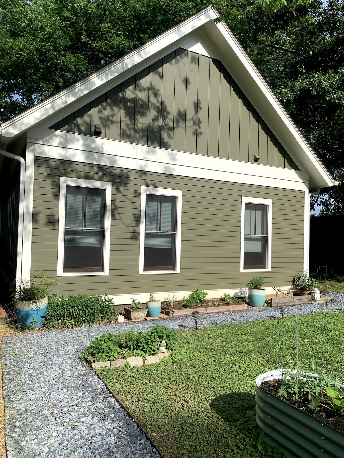 The exterior of the cottage is showcased, featuring a green facade and a gabled roof. Three windows are visible, framed by potted plants along a gravel path leading to the entrance, which is set in a well-kept yard with grass and garden beds.
