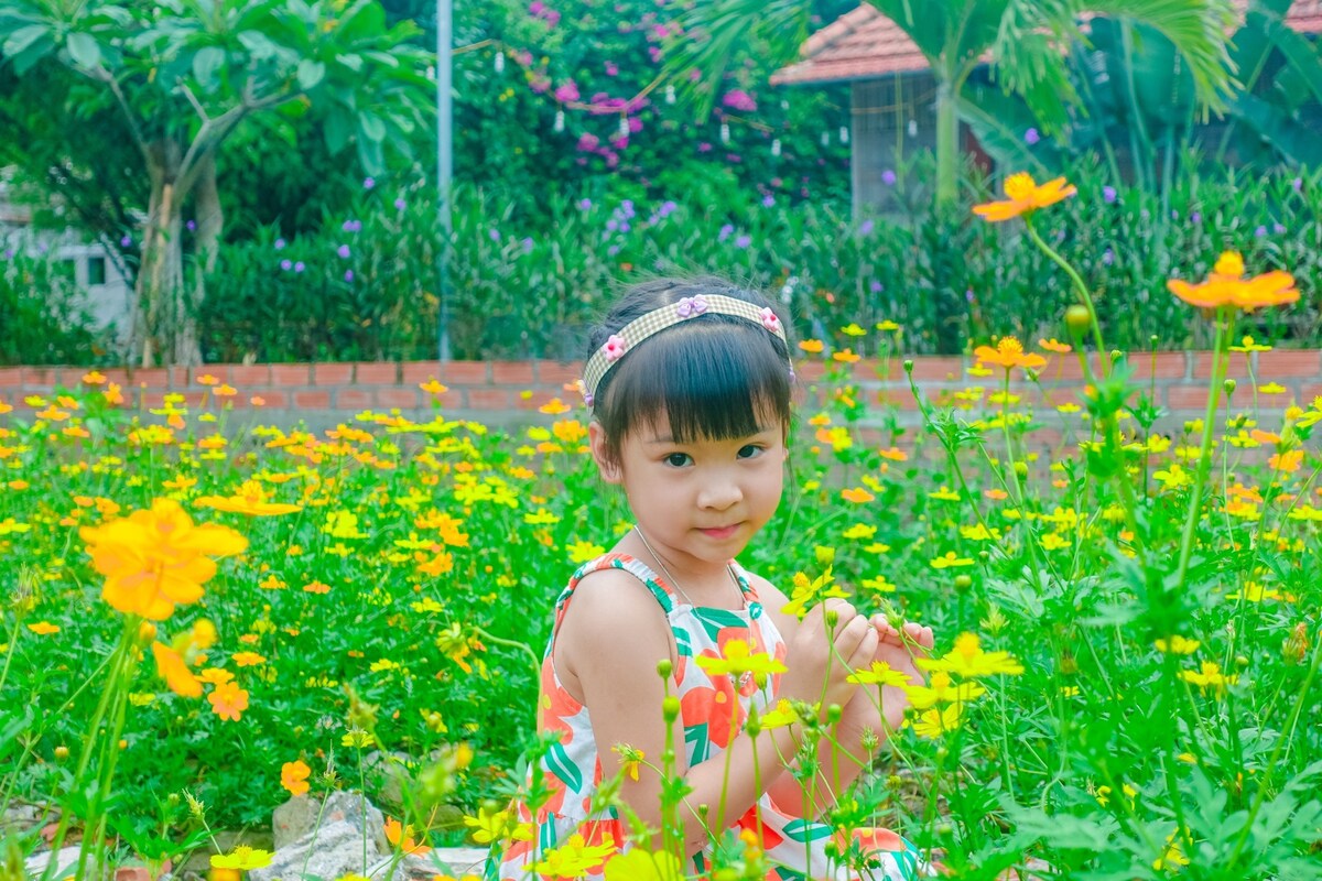 A lush garden filled with vibrant yellow flowers provides a colorful backdrop. A child dressed in a patterned dress is seated among the flowers, creating a serene and natural setting. Various green plants and trees are visible in the background, contributing to the garden's atmosphere.