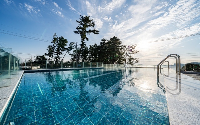 A tranquil swimming pool reflects the sky, surrounded by trees that frame the serene landscape. The pool features clear blue water with shimmering tiles, and glass railings provide an unobstructed view of the horizon.