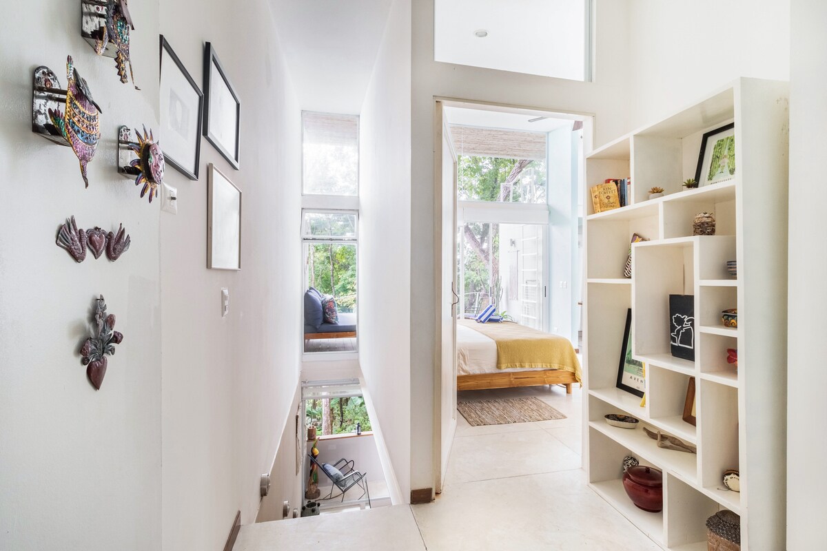 A well-lit hallway connects the living spaces, showcasing natural light filtering through large windows. Decorative local artworks are displayed on the walls, while shelves on the right exhibit an array of books and decorative items. A doorway at the end leads to a cozy bedroom area.