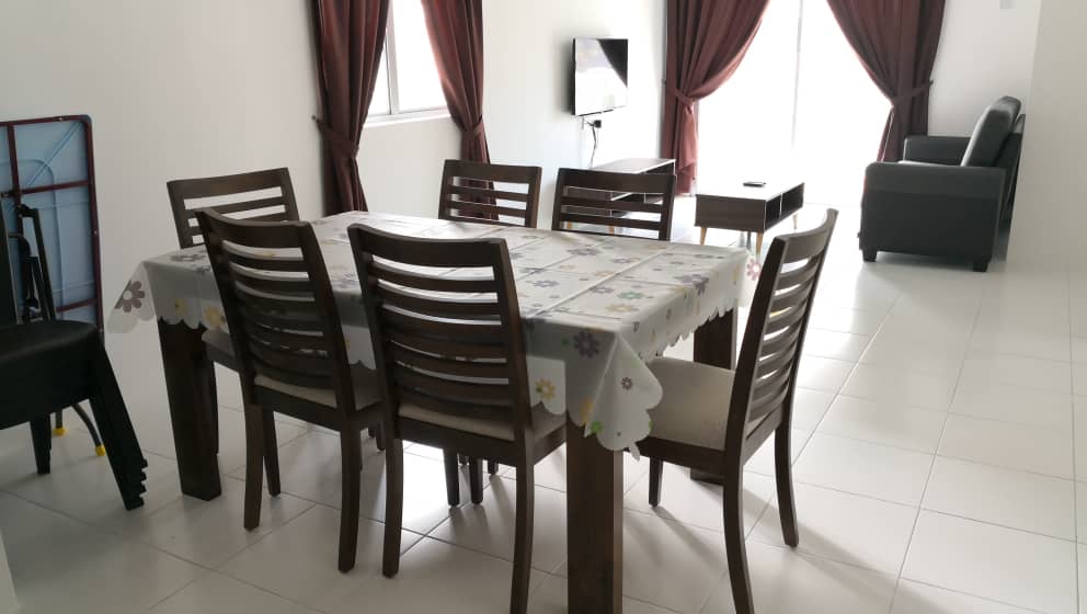 A dining area is depicted with a large wooden table surrounded by six chairs. A patterned tablecloth covers the table. Natural light is entering through large windows, which are draped with dark curtains. A flat-screen TV is mounted on the wall nearby.