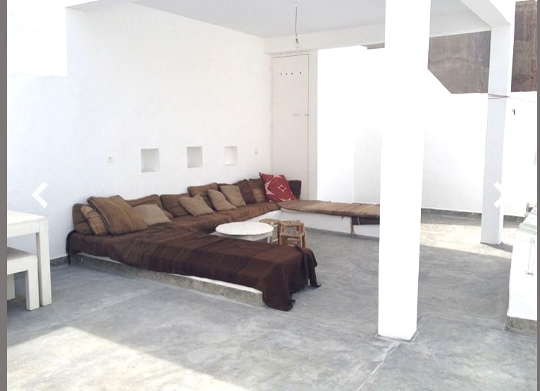 An indoor patio area features a spacious seating arrangement with inviting cushions and low tables. The white walls create a bright and open atmosphere, while the flooring is smooth and plain concrete. Natural light floods the space, enhancing its minimalist design.