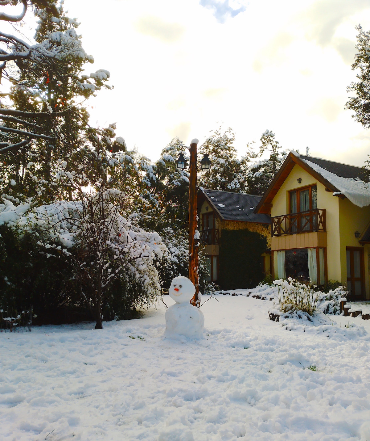 A snowy landscape surrounds a charming two-story home with wooden accents. A snowman stands playfully in the front yard, highlighting the winter scene. Trees and shrubs are coated in snow, creating a serene atmosphere amidst the white blanket covering the ground.