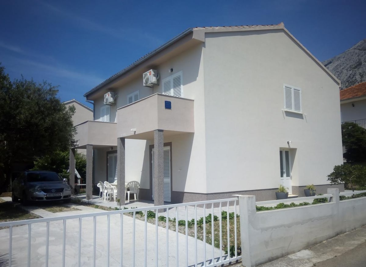 A two-story house is presented with a light-colored exterior, featuring two air conditioning units and white shutters. A small balcony is visible above the entrance, where two chairs are placed. A car is parked in the yard, and a low fence borders the property.