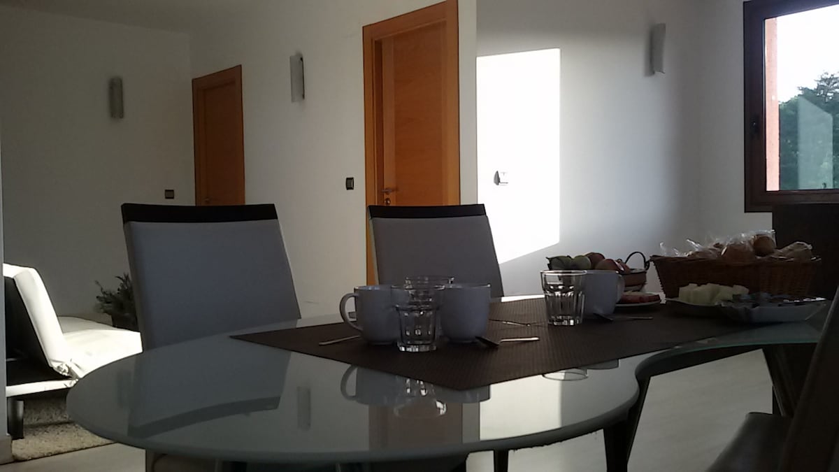 A modern dining area is visible, featuring a round glass table set with plates, cups, and a basket of fruit. The room is brightly lit by natural light coming through a nearby window, highlighting the clean lines and minimalist design of the space.