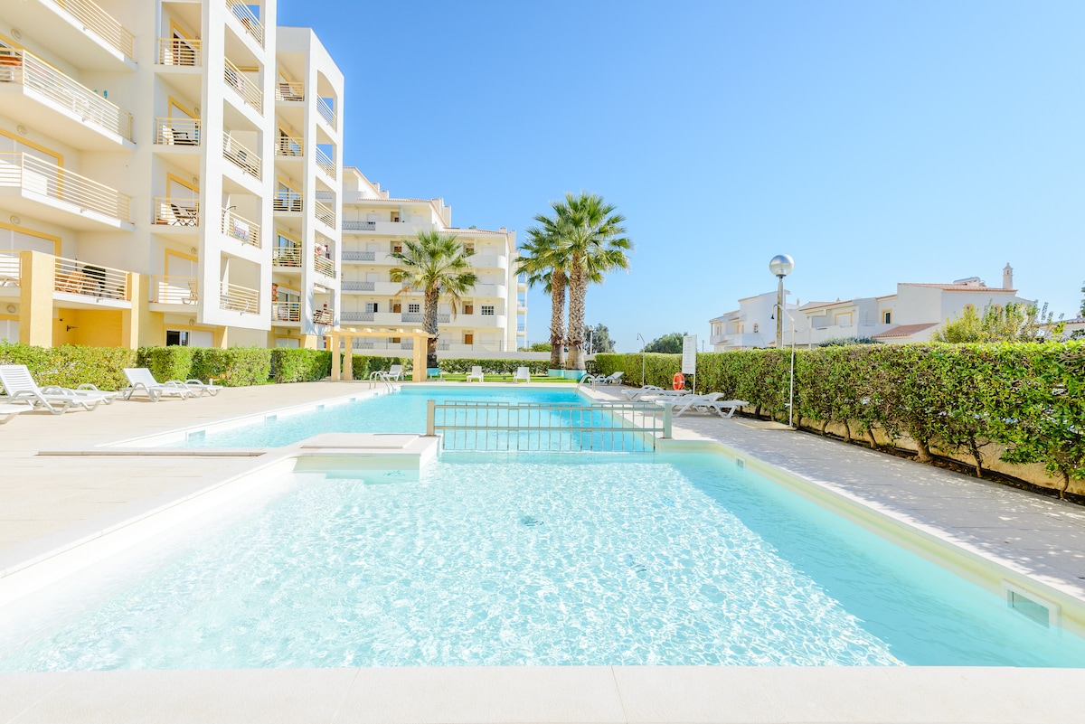 A clear blue swimming pool is surrounded by well-maintained green hedges and palm trees. The pool area features several lounge chairs and is situated adjacent to a modern residential building, providing a bright and inviting atmosphere under a clear sky.