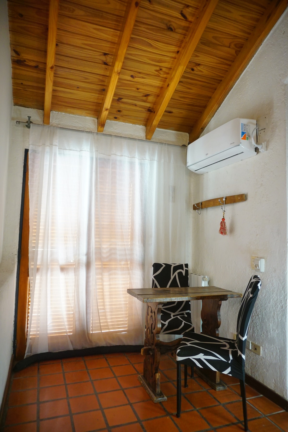 A small dining area is situated by a window, featuring a compact wooden table and two patterned chairs. Soft natural light filters through sheer curtains, illuminating the space. An air conditioning unit is mounted above for comfort.