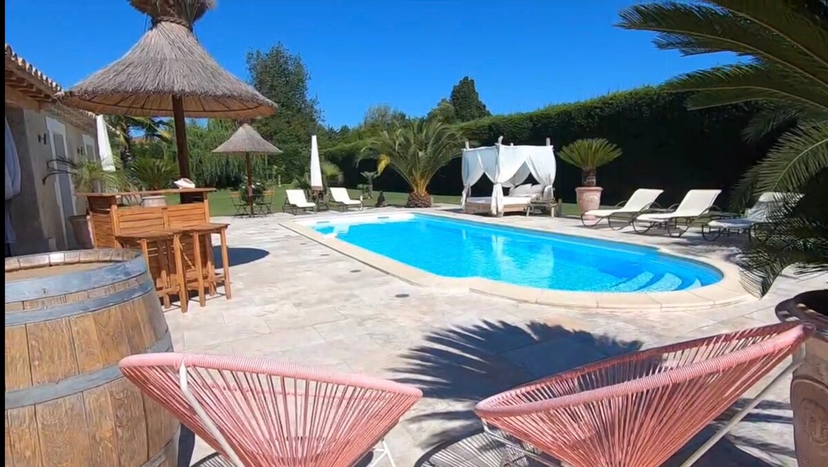 The outdoor area features a heated swimming pool surrounded by lounge chairs and palm trees. A thatched roof gazebo and a bar area enhance the space, complemented by a backdrop of well-maintained greenery and a clear blue sky.