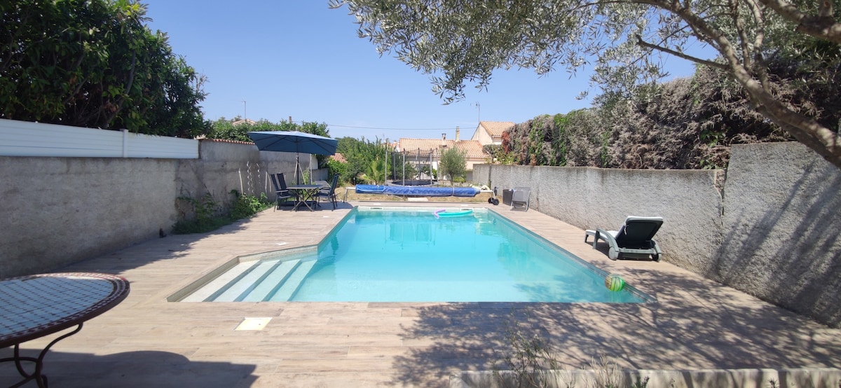 The outdoor area features a clear swimming pool surrounded by wooden decking. Sun loungers and shaded seating areas are visible, providing space to relax. Lush greenery lines the perimeter, enhancing the tranquil setting. A separate inflatable pool is located further away from the main pool.