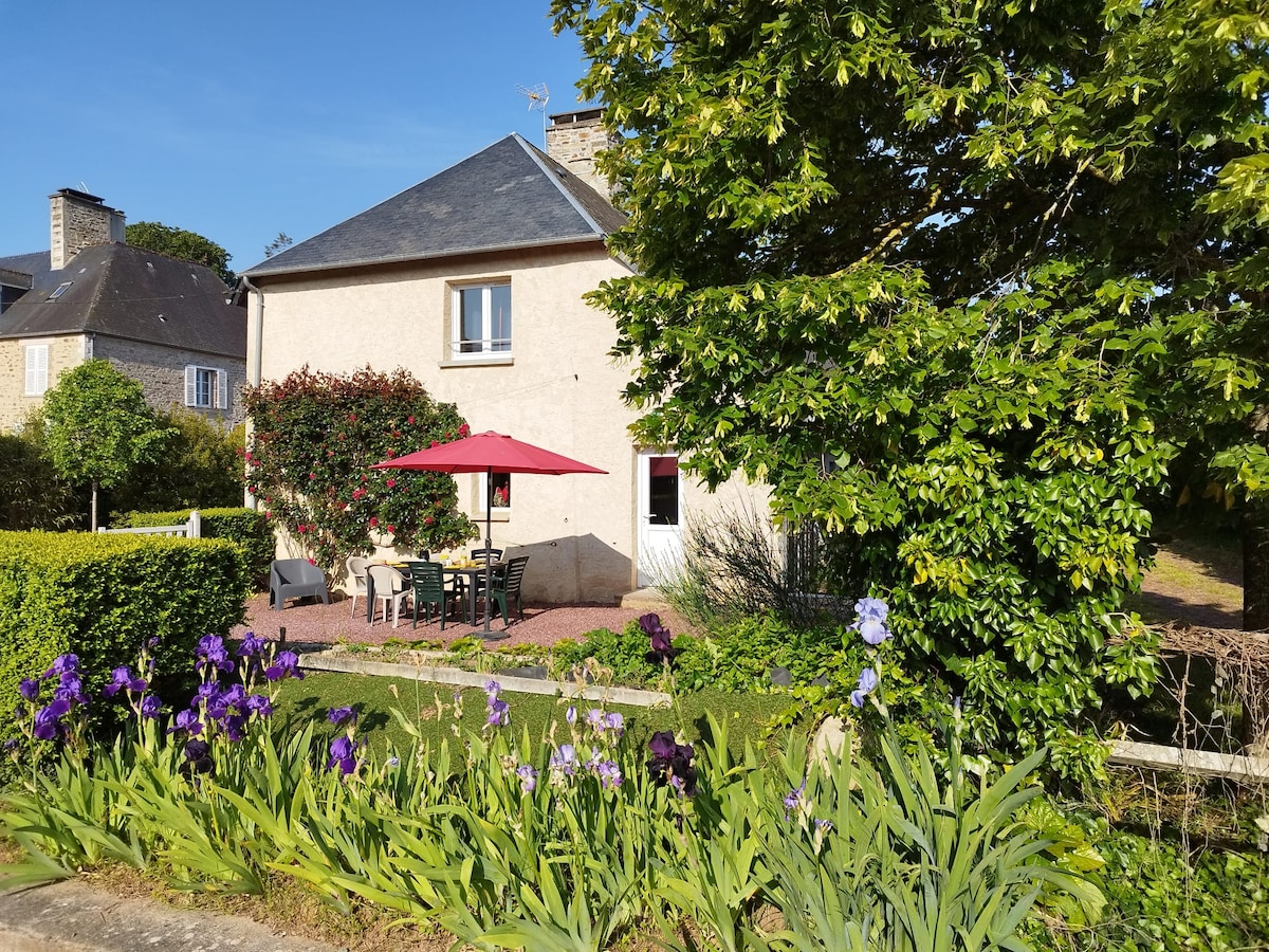 A charming farmhouse is surrounded by gardens featuring vibrant flowers and greenery. An outdoor dining area is shaded by a red umbrella. The building displays a neutral facade, with windows that complement the natural surroundings.