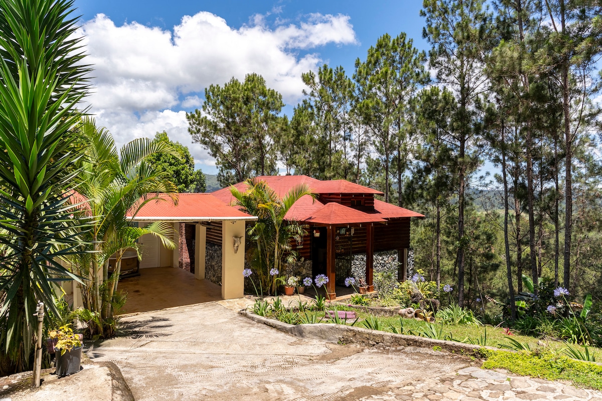 The house is set against a backdrop of lush greenery, surrounded by tall trees. A winding driveway leads to an inviting entrance, showcasing a red roof and stone accents. Colorful flowers and vibrant plants adorn the landscaping, enhancing the natural beauty of the area.