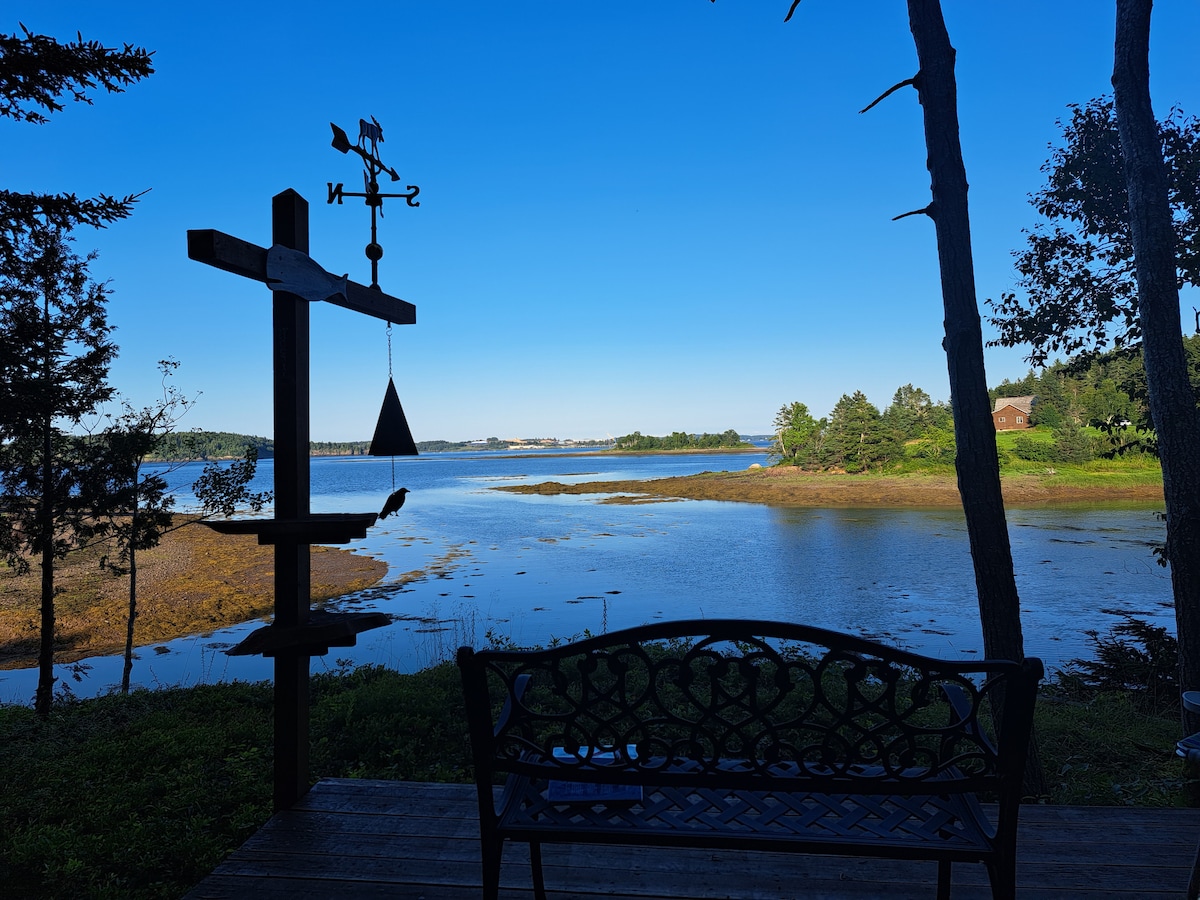 A serene view of Cobscook Bay is captured from a covered deck. A decorative weather vane is seen beside a bench, with calm waters and a clear blue sky serving as a backdrop. Lush greenery borders the scene, enhancing the natural setting.