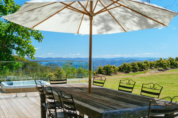 An outdoor patio with a wooden dining table under a large white umbrella overlooks a scenic landscape of rolling hills and orchards. A jacuzzi sits nearby, with clear glass fencing separating the patio from the expansive green lawn.
