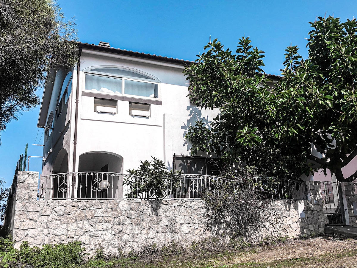 The exterior of a two-story house is visible, showcasing a stone wall at the base and large windows on the upper level. Lush greenery surrounds the building, with glimpses of a covered porch area. A clear blue sky provides a bright backdrop.