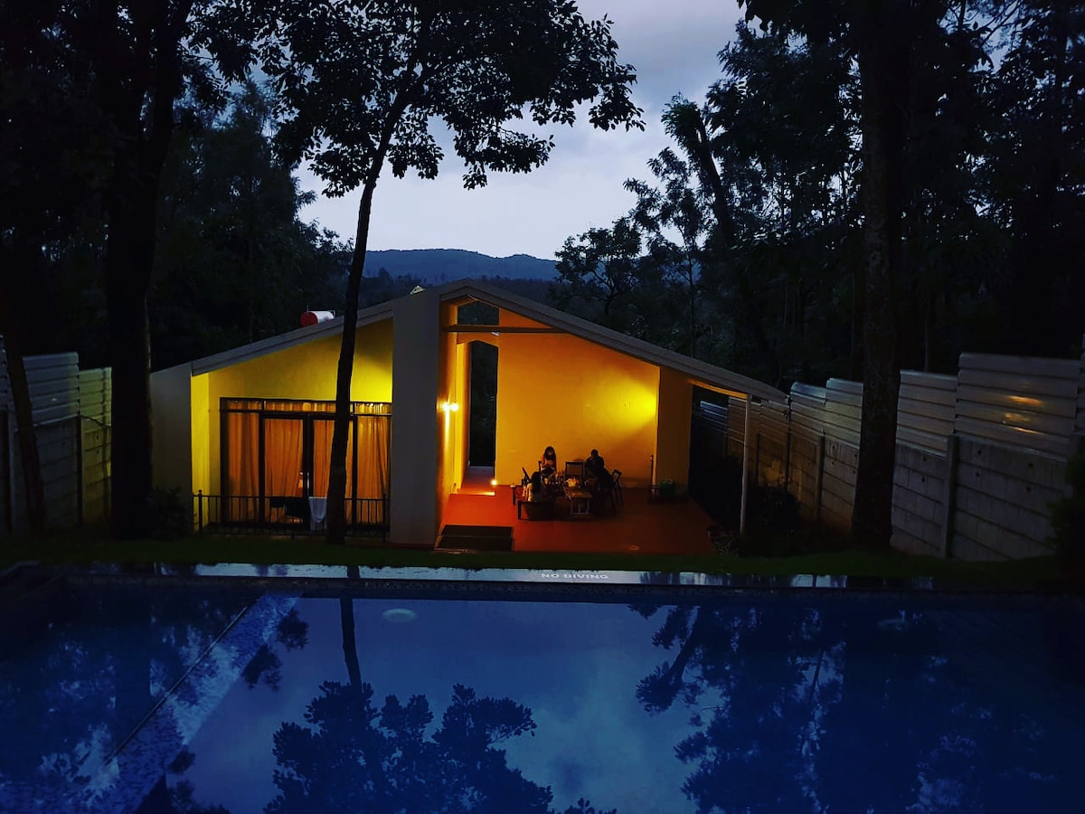 A modern villa is set against a twilight backdrop, featuring large glass windows that reflect the soft glow from inside. A tranquil pool mirrors the surrounding trees, while a cozy outdoor seating area is visible on the patio, inviting relaxation amidst nature.