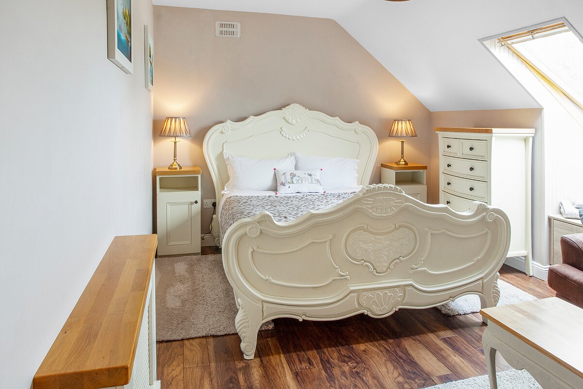 A well-appointed bedroom features a spacious white bed with decorative detailing, complemented by soft lighting from bedside lamps. Light wood furniture, including a dresser and side tables, adds a touch of elegance, while a window with a sloped ceiling allows natural light to fill the room.